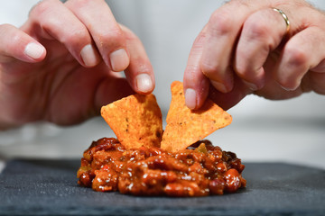 Close up of male hands putting nachos on chili on a dark plate. Exotic food and cooking concept.