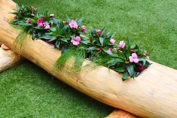 Pink flowers in big natural wooden pot made of tree trunk. Flowerbed with blooming flowers in retro planters on green artificial grass cover outdoors in sunny summer day.
