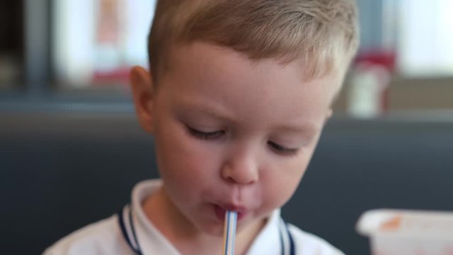 Cute blond caucasian boy portrait drinks sweet soda through tube and puts white paper cup on table in fastfood restaurant. Quick snack in fast food, quench your thirst with cold soda.