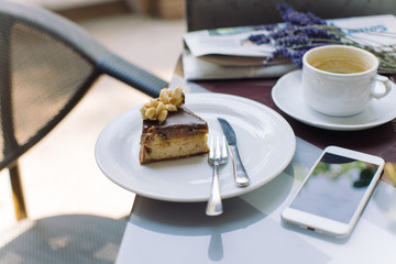 Cheesecake sliced and plated, cup of cappuccino, smart phone on table. Coffee break on summer terrace.
