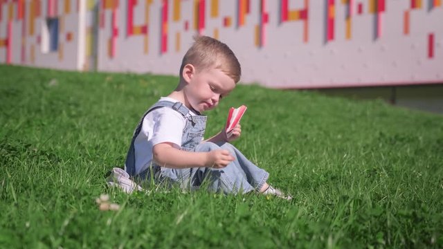 Blond Caucasian boy sits on grass in summer weather outside and eats fried French fries, child puts potatoes in red barbecue sauce and puts them in his mouth. Fast food and snacks in nature.