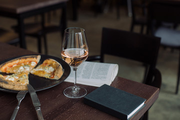 Glass of rose wine, 4 cheese pizza, newspaper and notebook on wooden table at the cafe. Day off meal outside home.