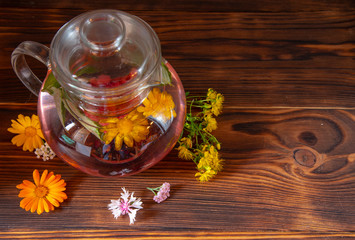 A transparent teapot with herbal tea and medicinal plants on a dark wooden table top.