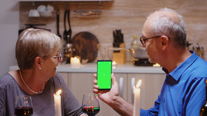 Old senior retired couple holding green screen phone at dinner. Aged people looking at mockup...