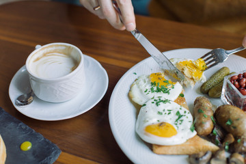 Close up of man's hands holding fork and knife while eating traditional English breakfast with fried eggs, sausages, beans, mushrooms, bacon. Lifestyle concept.