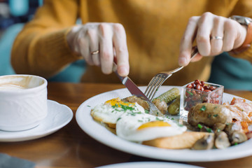 Man eating traditional English breakfast with fried eggs, sausages, beans, mushrooms, bacon at cafe in the morning.