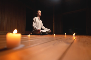 A portrait of an attractive Caucasian man sits in cotton robes in a lotus pose in a dark wooden...