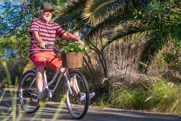 Cheerful senior man with white beard riding with a vintage bicycle enjoying freedom, basket with daisies - concept of active playful elderly during vacation