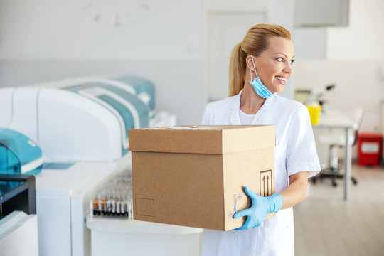 Smiling blond female lab assistant standing and holding box with medicines and vaccine.