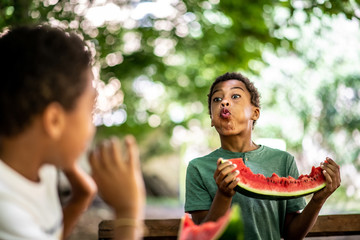 Two boys are sitting at a table eating a watermelon, one is spitting at the other with a watermelon...
