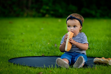 A little pretty baby sits on the lawn in the garden and eats a fresh baguette.