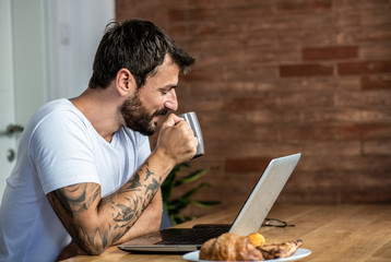 A man is sitting at home, drinking coffee and preparing to work on a laptop