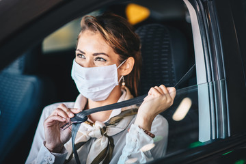 Fototapeta na wymiar A young woman sits in a car with a mask on her face and adjusts her seat belt, life during a virus pandemic