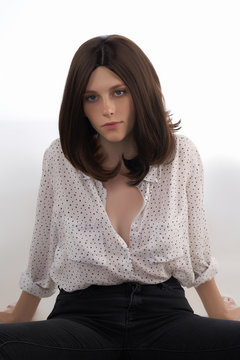Young woman in a natural looking dark brown wig without bangs is sitting on the gray backdrop. The lady in shoulder-long wig is wearing black jeans and a white unbuttoned shirt with polka-dot print. 