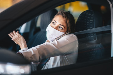 Fototapeta na wymiar A young woman drives a car with a mask on her face, holds a mobile phone with her shoulder while driving, life during a virus pandemic