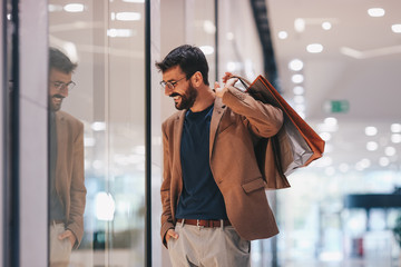 A man walks through a shopping mall with a smile on his face and bags in his hand and looks at the shop windows