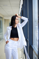 Fototapeta na wymiar Young brunette woman, wearing white pants and jacket, standing in light passageway, posing for social media, leaning on window. Businesswoman on a break. Female portrait photography.