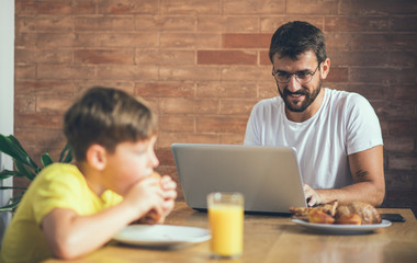 A father and son sit in the kitchen during the morning, a father works on a laptop and his son has breakfast