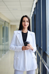 Young brunette woman, wearing white business suit, holding black paper coffee cup, standing in light office building near window. Businesswoman on lunch break. Corporate culture concept.