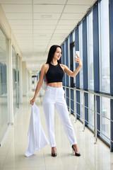 Young brunette woman, wearing white pants and black top, holding jacket in hand, taking selfie picture with cell phone in light passageway, posing for social media. Businesswoman on lunch break.