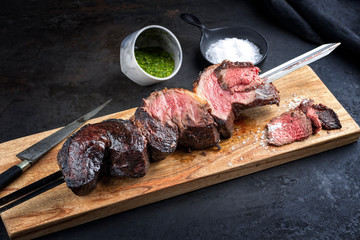 Barbecue dry aged wagyu Brazilian picanha from the sirloin cap of rump beef offered with chimichurri sauce as closeup on a skewer on a wooden design board on rustic black background