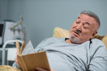 A handsome senior man reading a book in bed at the hospital ward