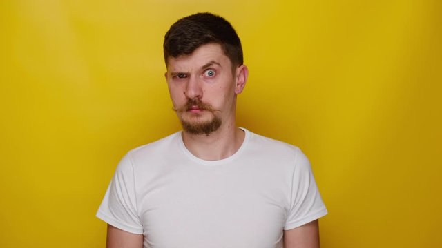 Portrait of funny attractive young man frowning his face raising up eyebrows, looking shocked at camera, dressed in white t-shirt, isolated on yellow studio background with copy space