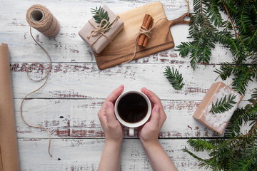 hands hold white cup of coffee on an old wooden work table with handmade gifts and branches of Christmas tree.Flat Lay.Top view