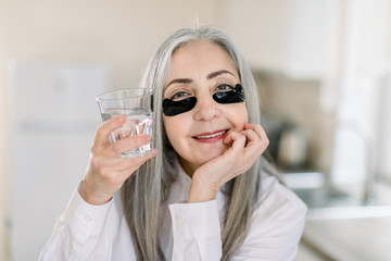 Elegant retired woman with long gray hair doing cosmetics procedures, posing to camera with black eye patches under eyes and glass with water. Beauty treatment at home kitchen interior