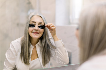 Facial mask and anti wrinkle therapy. Smiling elderly woman standing in front of bathroom mirror and remowing black eye patches after getting her morning spa procedures