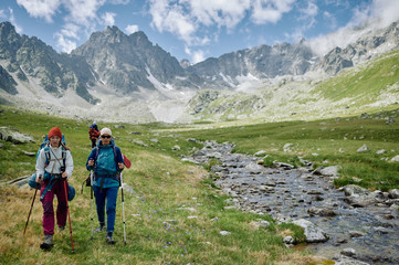 Fototapeta na wymiar Two woman hikers are walking with trakking poles by the river among mountains. Extreme tourists in wild nature. Domestic travel and trekking. Local tourism. Healthy lifestyle