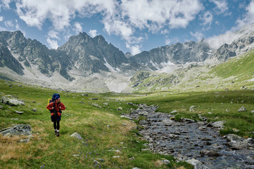 Fototapeta na wymiar Alone hiker man with backpack is walking by the river among mountains. Extreme tourist in wild nature. Domestic travel and trekking. Local tourism. Healthy lifestyle