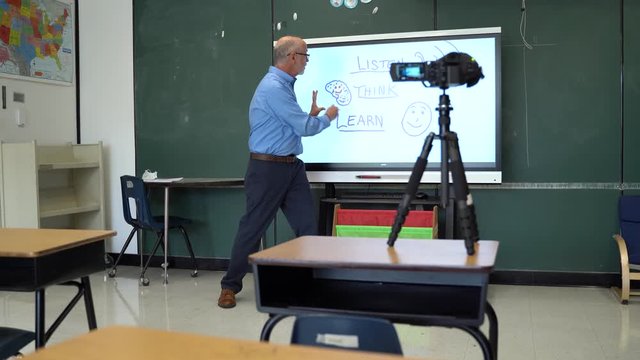 Male teacher using an interactive whiteboard in an empty classroom teaching to a video camera.