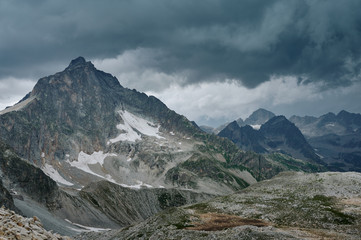 Gloomy mountain landscape with thunder cloudy sky, rocky ranges and peaks with glaciers and snow fields. Wild nature valley