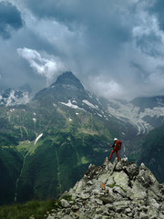 Obraz premium Alone hiker man with backpack and helmet against the gloomy mountain peak landscape with thunder cloudy sky, rocky ranges and peaks with glaciers. Domestic travel and trekking. Local tourism.
