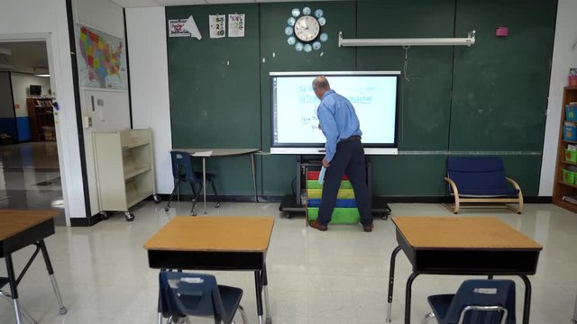 Camera pushes in on a male teacher writing on an interactive whiteboard in an empty classroom.