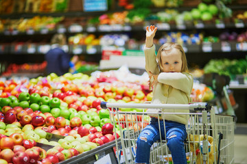Adorable toddler girl sitting in the shopping cart in a food store or a supermarket