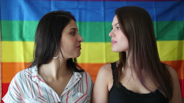 Lesbian girlfriends portrait smiling at camera with rainbow flah in background. LGBT community