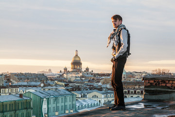 A bearded guy in a leather vest and jeans stands on the edge of the roof with a view of St....