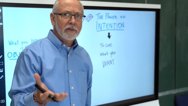 Closeup portrait of happy teacher wearing glasses and talking to camera in front of interactive whiteboard.