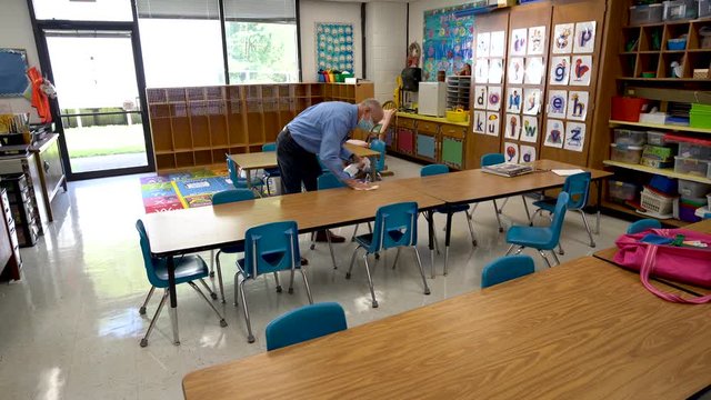 Wearing a medical face mask, a male teacher disinfects chairs and table in an empty classroom during covid-19.