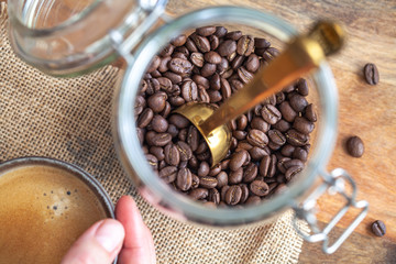 Top down view of a jar of coffee beans with a measuring spoon and a person holding a cup of coffee