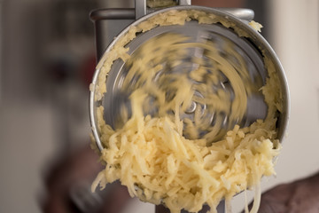 Detail einer drehenden Kartoffelreibe und geraspelten Kartoffeln mit Händen im Hintergrund, die...