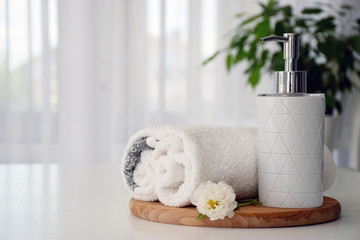 Fototapeta na wymiar Fresh white towels folded on wooden board, white flower and liquid soap container with green leaves of house plant and tulle window on background. Spa and wellness or beauty salon concept. Copy space.