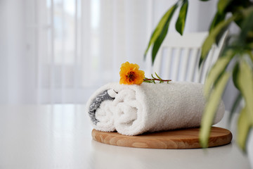 Fresh white towel rolled on wooden board, orange flower and tulle window on background. Hotel spa and wellness concept. Copy space.