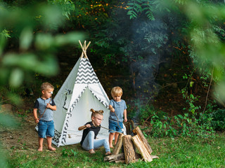 Three young kids roast marshmallows in the backyard in front of a play teepee © Drew Woolery