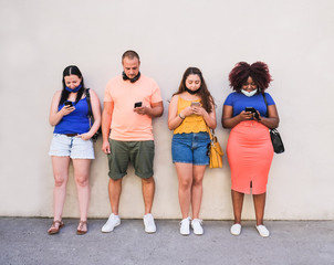 Obraz premium Young multiracial people standing in a line and looking at their mobile phones while wearing safety face mask - Coronavirus lifestyle