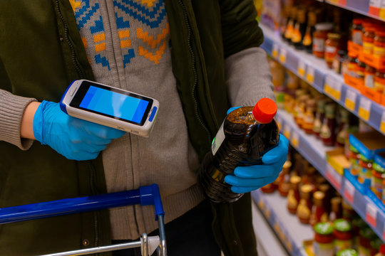 A customer wearing medical protective gloves holds a barcode scanner and scans the product. Cropped shot, no face visible. Self-service in the store.