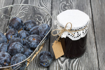Jar of plum jam. With an inscription tag. Nearby plums in a basket. On painted pine planks.