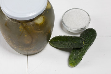 Jar of pickled cucumbers. Nearby are fresh cucumbers and a container of salt. On a painted board surface.
